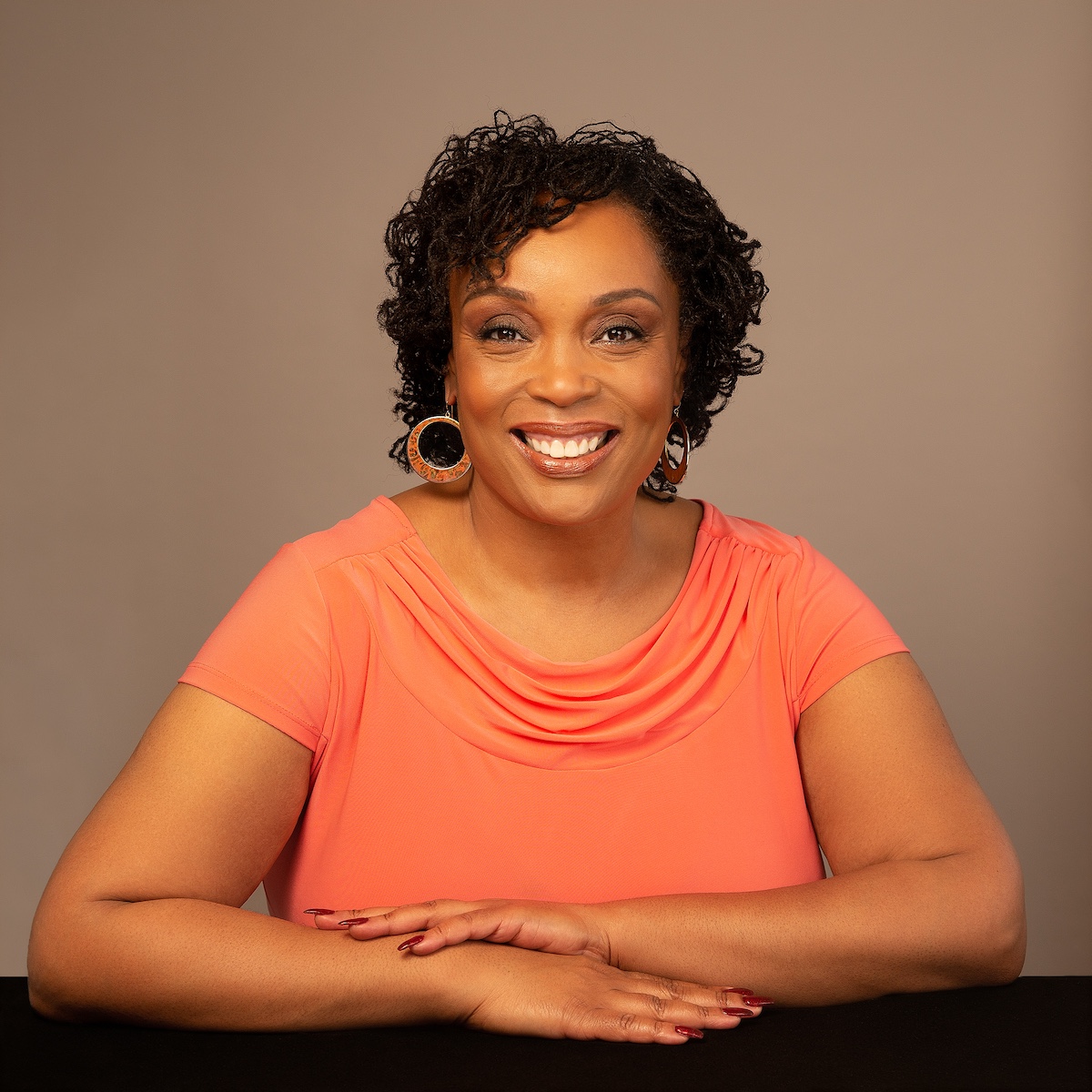 Dr. Renai Ellison headshot with an orange shirt and black table.